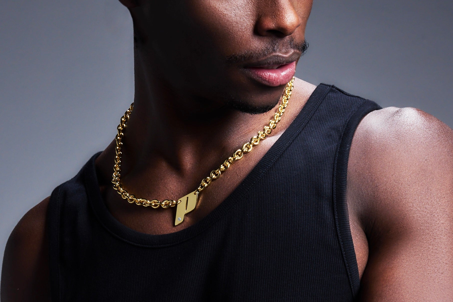 Close-up of male model wearing Play’s 18K gold monogram pendant necklace on a chunky chain, styled with a black sleeveless top against a gradient backdrop. Banner image for the necklaces and pendants from the OTR: On the Run collection.