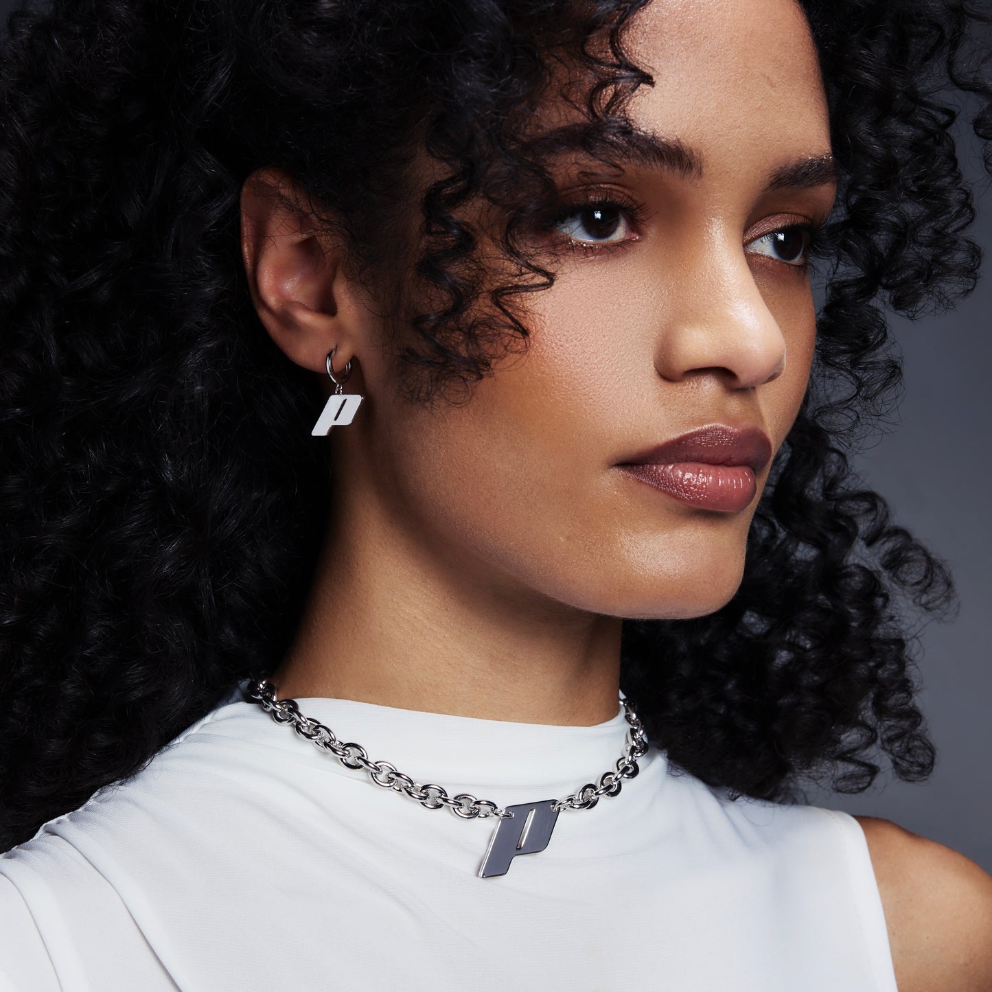 Model wearing Play's sterling silver monogram earrings and matching pendant necklace, styled with a white high-neck top against a gradient backdrop.