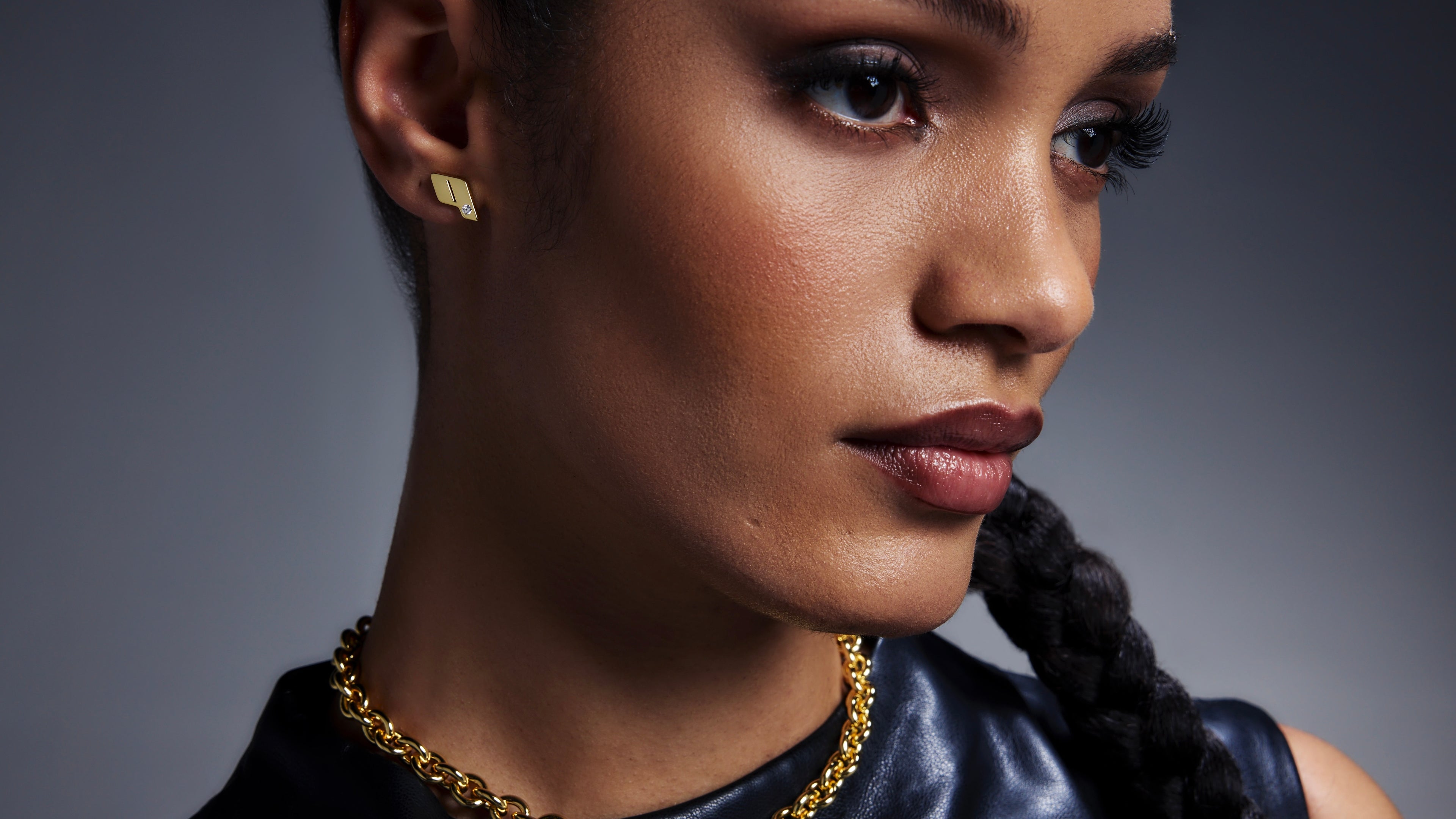 Close-up of model wearing Play stud earrings and the 18k Play monogram pendant necklace, shot against a dark gradient backdrop.