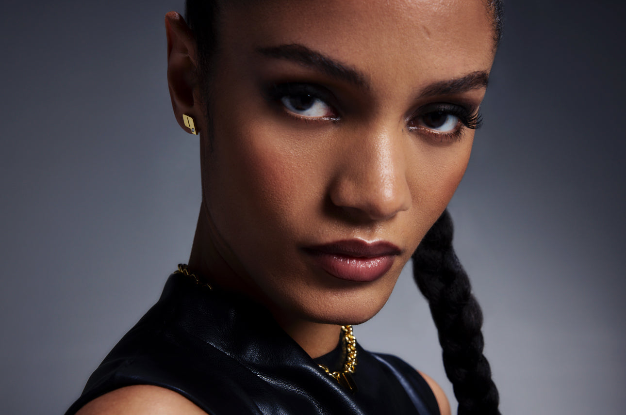 Close-up of model wearing Play’s 18K gold monogram earring and matching chain necklace, set against a soft gradient backdrop. Banner image for the OTR: On the Run collection.
