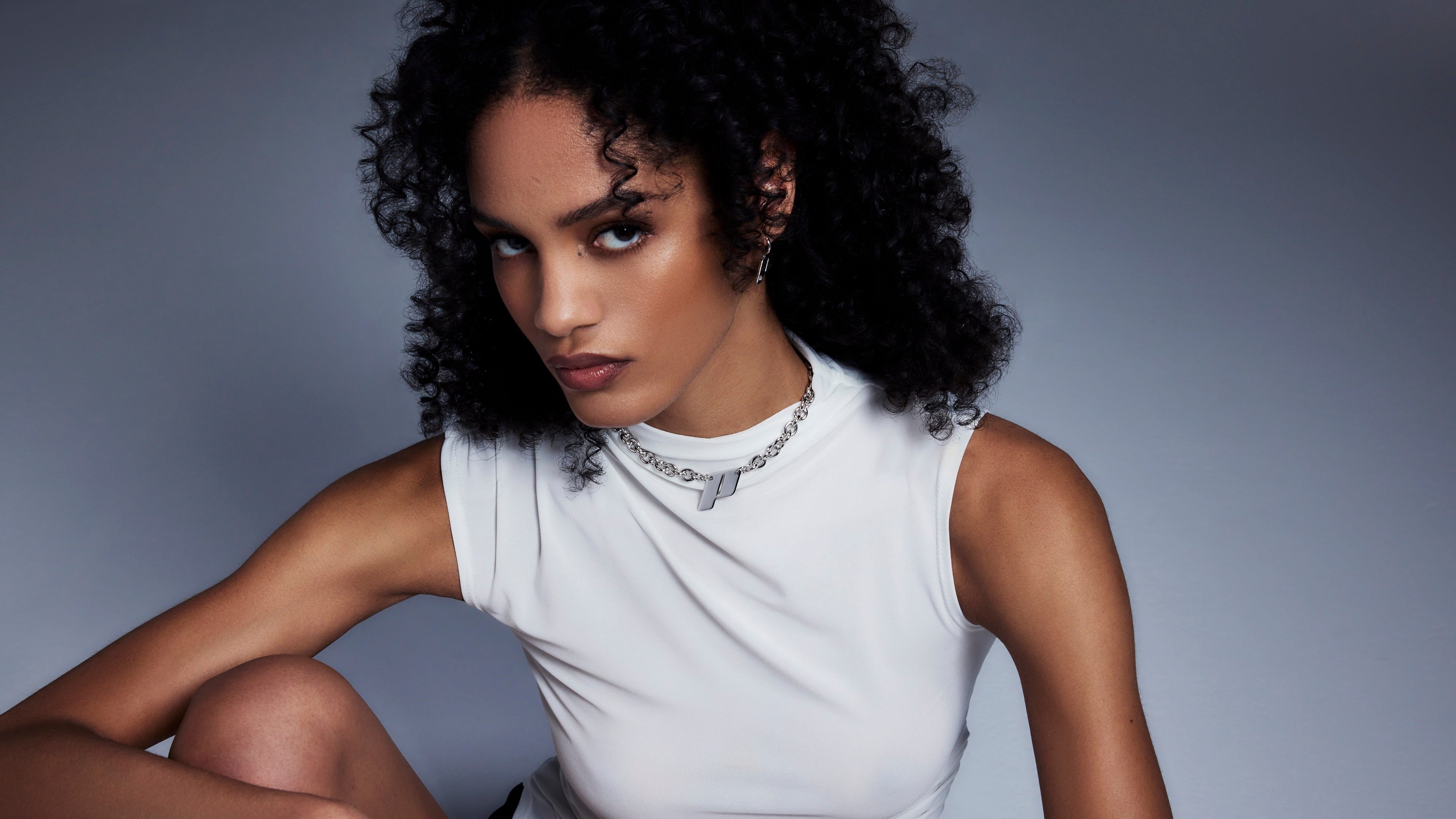 Close-up of a female model with curly hair wearing Play’s sterling silver monogram necklace and earrings. Styled with a white sleeveless top against a soft gradient backdro