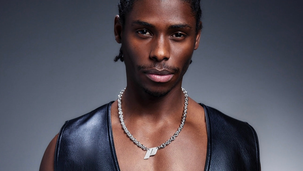 Portrait of male model wearing Play’s sterling silver monogram necklace on a bold chain, styled with a black leather vest against a soft gradient background. Banner image for the OTR: On the Run sterling silver necklace edit.