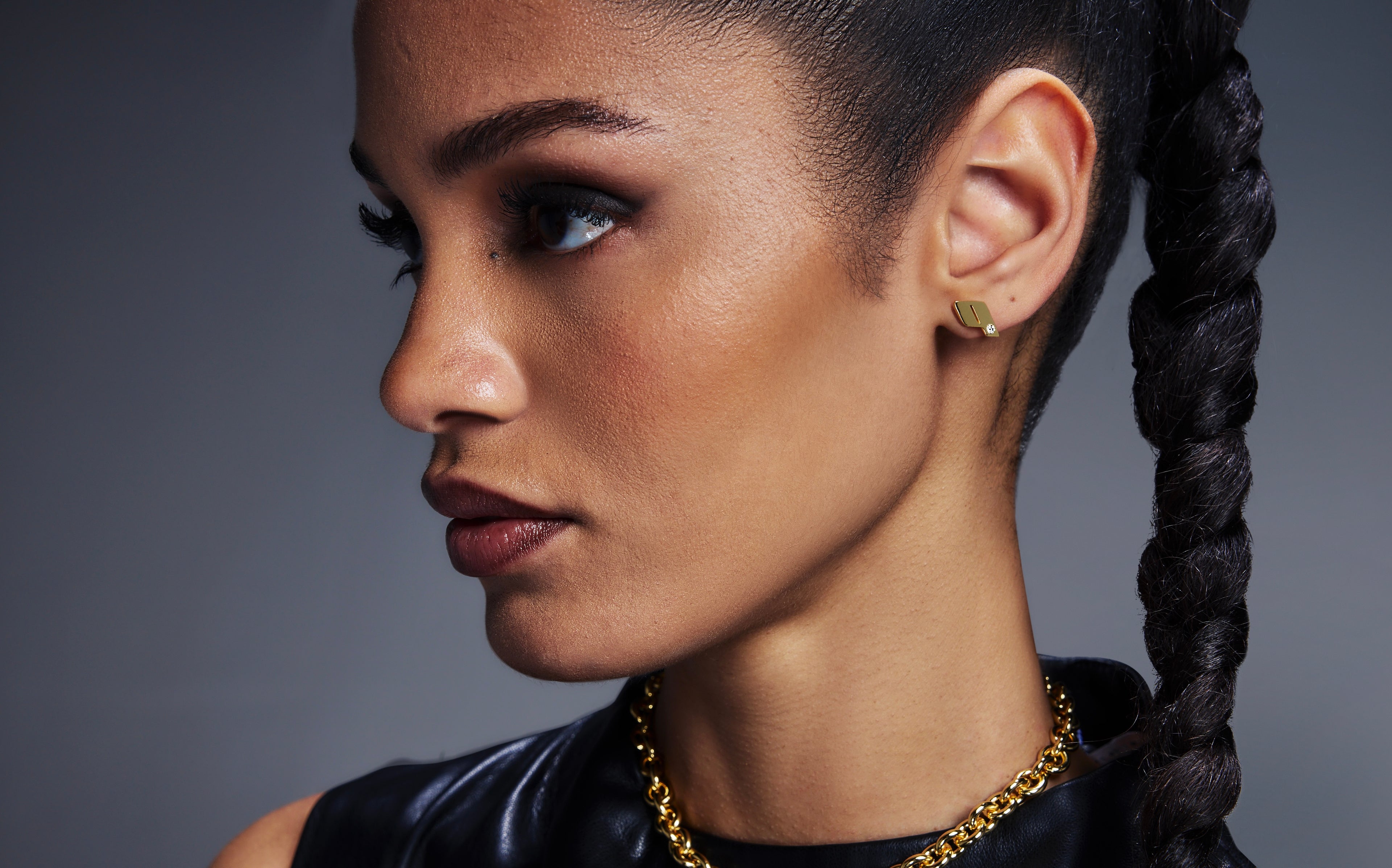 Side profile of a model wearing Play's 18k yellow gold monogram stud earring and matching pendant necklace, styled with a sleek braid.