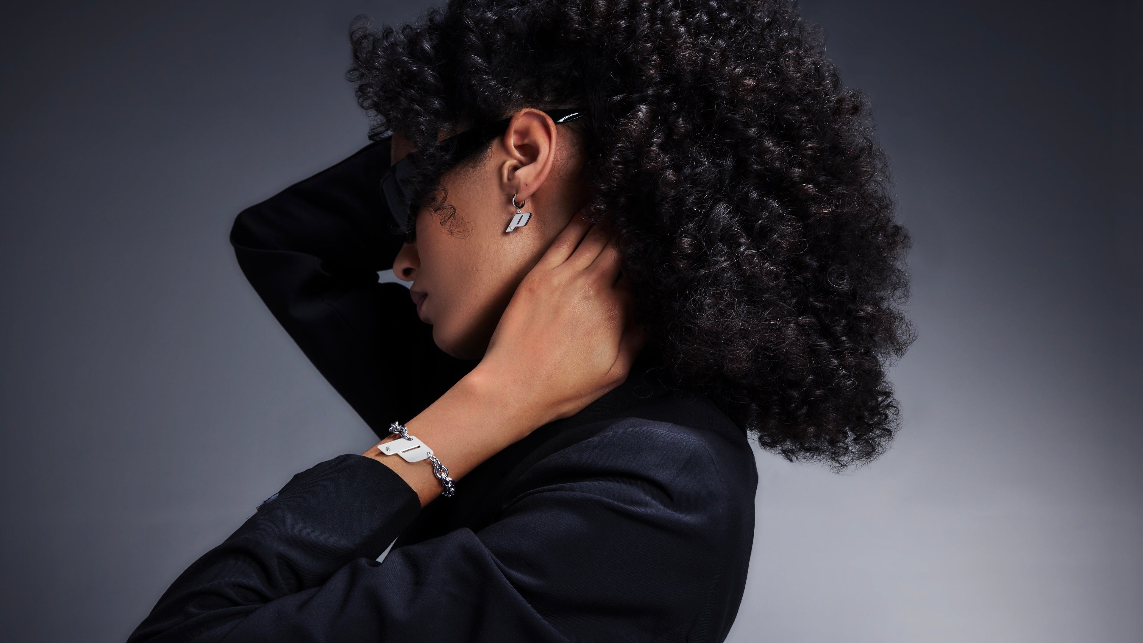 Side profile of a female model wearing Play’s sterling silver monogram earrings and matching bracelet. Shot against a soft gradient backdrop, styled with dark sunglasses and a black blazer.